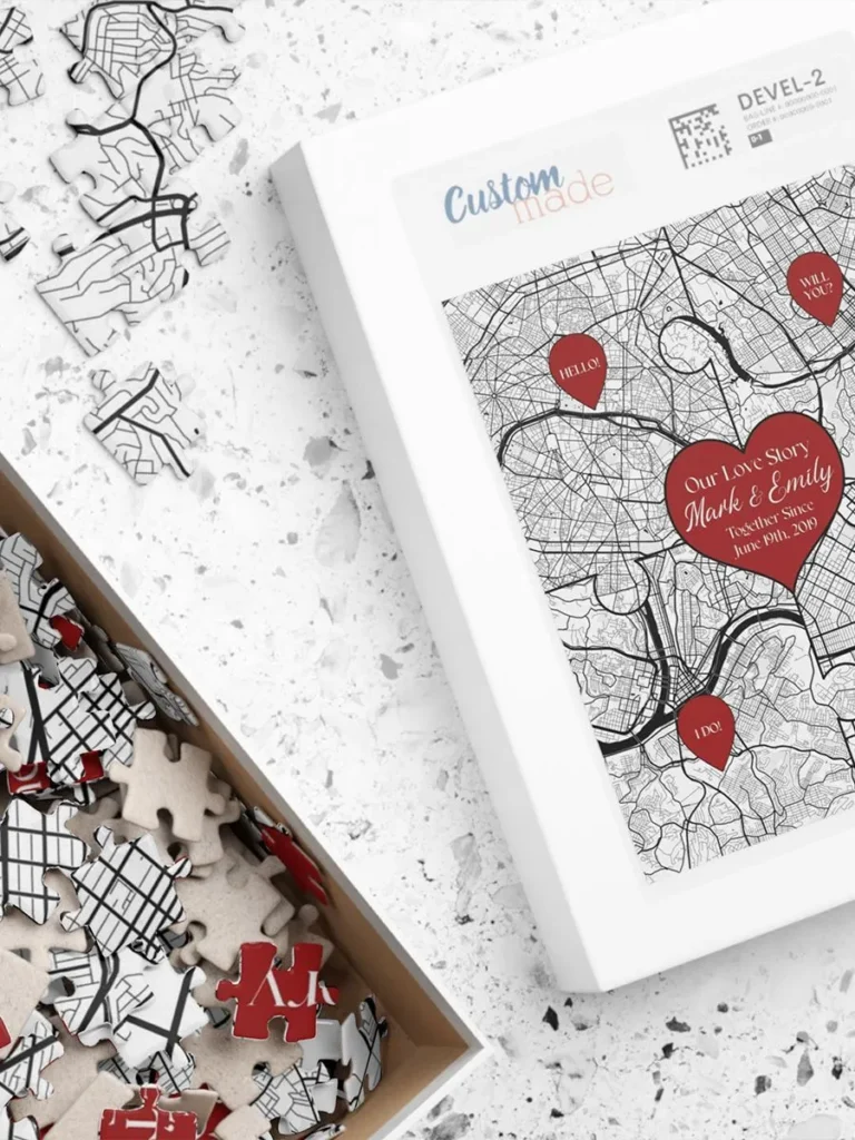 Custom love story jigsaw puzzle design, made up of four personalized maps and featuring two names and date within heart symbol, displayed on table with loose puzzle pieces inside and outside open puzzle box.